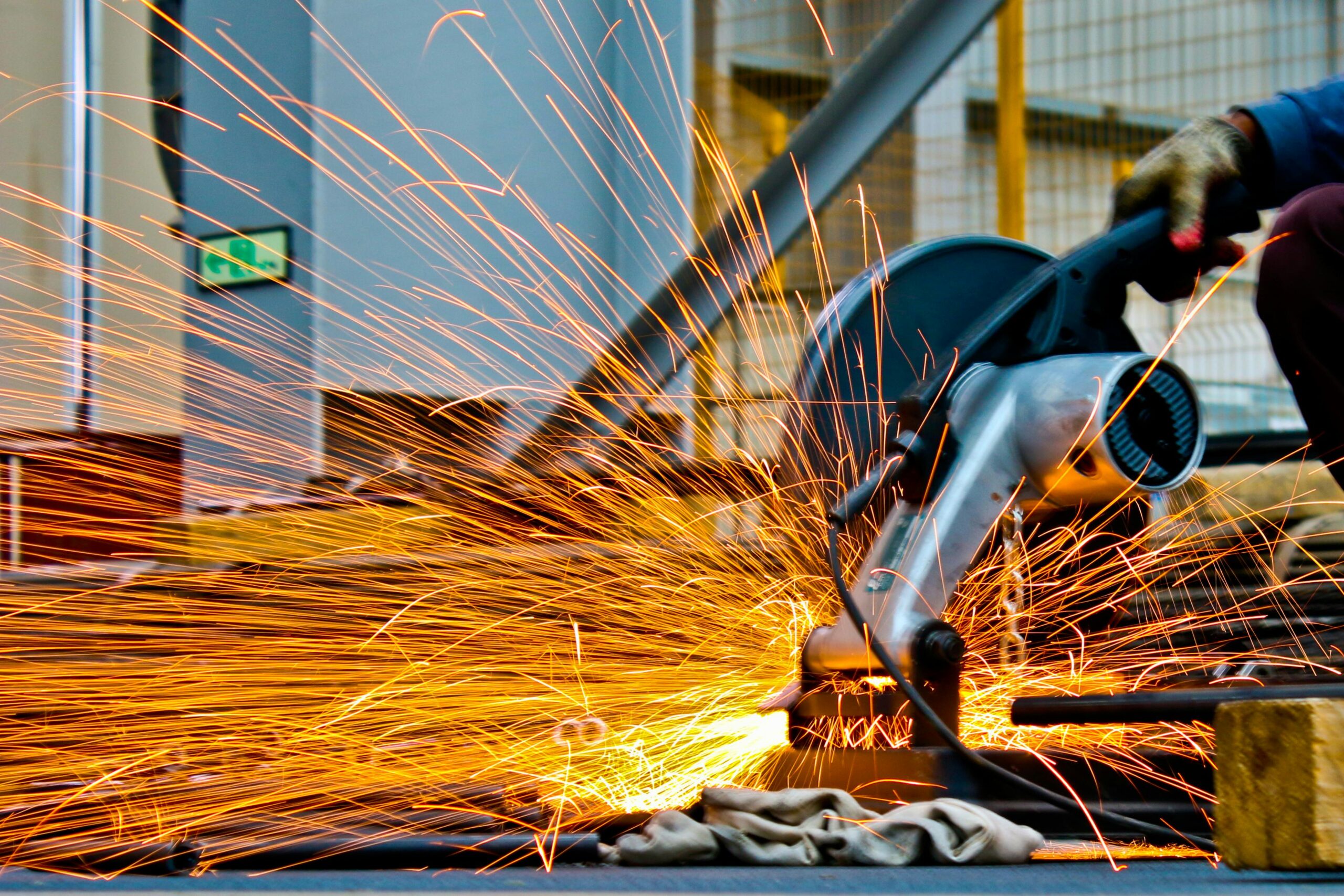 A close-up of a worker using an electric metal-cutting saw, producing a large spray of bright orange sparks in an industrial setting. The worker wears a protective glove, and safety signage is visible in the background.